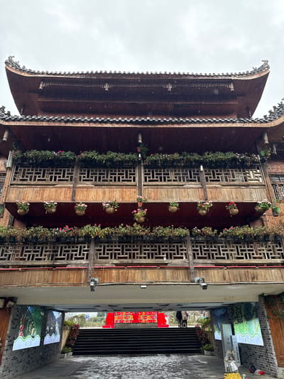 This image captures a multi-story building with traditional Chinese architectural elements in Gongtan, China, under an overcast sky with visible precipitation.

The building features a dark brown wooden facade with three prominent levels above a ground-level archway. The uppermost section displays curved, dark tiled roofs. Each visible upper level incorporates a balcony with intricate wooden lattice railings. The railings are adorned with rows of potted plants, primarily green foliage, and several hanging baskets containing pink and red flowering plants.

At ground level, a wide concrete archway serves as a passage. The paved surface beneath the archway appears wet. On the left interior wall of the archway, two vertical screens display green and white graphics. Inside the archway, a set of dark, wide stairs ascends upwards and outwards. Beyond the stairs, a brightly lit red and gold structure or banner is visible. Two individuals, dressed in coats, stand near the top of these stairs, to the right of the central axis. In the distance, beyond the red structure, a body of water and faint structures are discernible under the cloudy sky.