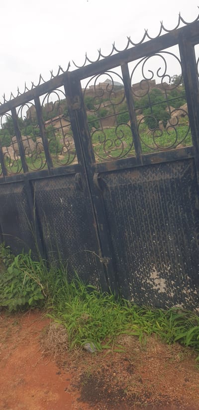 An outdoor, low-angle shot captures a weathered, black metal gate with decorative scrollwork in the upper section and a diamond plate pattern in the lower panels. The gate is partially obscured by green grass and dry vegetation growing in the foreground. Beyond the gate, a rocky, hilly landscape is visible with scattered trees and some simple structures. The sky is overcast and a pale gray. The image appears to be taken in Barikin Jirga Summa, Nigeria, given the context.