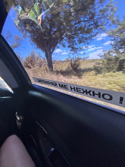 The image shows a view from inside a vehicle, looking out the passenger side window. A white sticker with black Russian text is affixed to the bottom of the window. The text reads "ЗАВТРАЙ МЕ НЕЖНО !" which translates to "Tomorrow, gently!". Outside the window, a tree is visible against a bright blue sky with scattered clouds. The landscape beyond appears to be a grassy slope with bushes and trees, and a white building is visible in the distance. The interior of the car is dark, with the armrest and part of a person's leg visible in the foreground. The scene suggests a journey through a rural or semi-rural area, possibly during the day. There is no specific visual information in the image that allows for the determination of its location in Novi Pazar, Bulgaria.