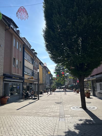 The image depicts a sunlit pedestrian street in the city of Heilbronn, Germany, on a clear day. A large, mature tree dominates the right side of the frame, casting a long shadow across the patterned paving stones. Along the left side of the street, buildings house various businesses, including a "Prince Barber" with its interior visible, and a shop labeled "MR. BAKER". Several people are scattered along the street, some seated at tables outside a cafe. Colorful, decorative lanterns hang suspended above the street, adding a festive touch. The scene is one of urban activity and everyday life, with a focus on the pedestrian environment and the architecture of the surrounding buildings.