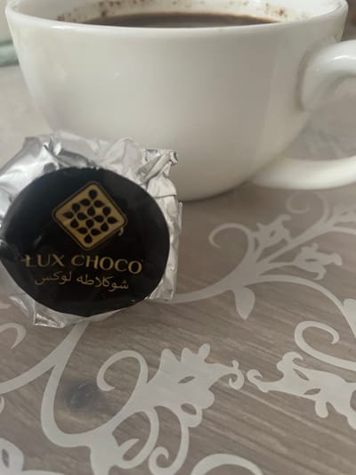 A close-up shot shows a white mug of coffee and a wrapped chocolate. The mug is on the left side of the frame, and its contents are dark brown. Next to the mug, on the right, is a round, dark chocolate wrapped in silver foil. The chocolate's wrapper is partially open, revealing a black circular label. The label features a gold border and the text "LUX CHOCO" in white. Below "LUX CHOCO" is Arabic text in gold. Inside the gold border is a stylized image of a chocolate bar with small dots representing chocolate chips. The background is a patterned surface with white floral designs against a light brown wood-grain texture. The lighting appears to be natural daylight, suggesting it might be morning or afternoon. The scene evokes a sense of a quiet moment, perhaps a coffee break. The location is Brovary, Ukraine.