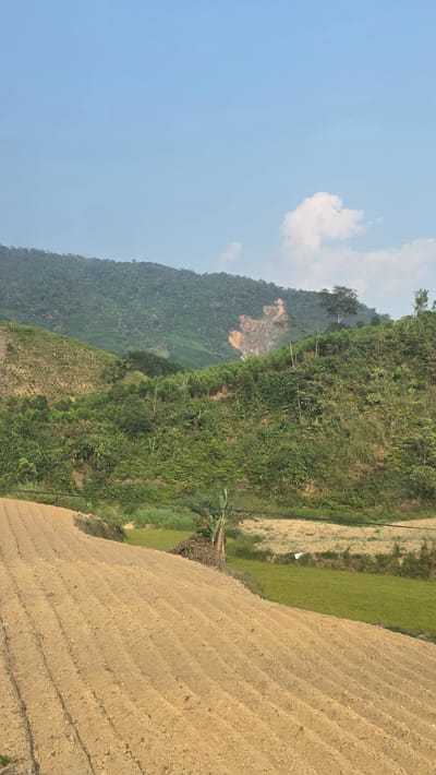 The image shows a rural landscape in Thôn 18, Vietnam. Terraced farmland, prepared for planting, dominates the foreground. A mountain range covered in lush green vegetation rises in the background. A patch of exposed earth is visible on one of the mountains. The sky is blue with a few white clouds. A small banana plant is near the cultivated land. Some green rice paddies lie in between the cultivated land and the vegetation.
