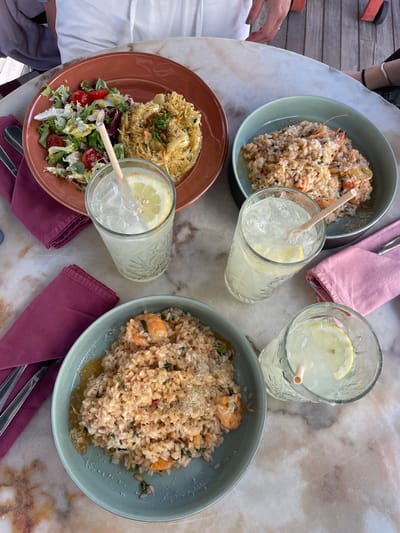 A high-angle shot shows a dining table setting, likely outdoors due to visible wood decking in the background. The table is a light-colored marble with noticeable veining patterns. Four dishes are arranged around the table: two appear to be shrimp risotto in blue-green bowls, one is a salad topped with a pasta-like dish on a reddish-brown plate, and the last looks like a rice dish. Each dish has a corresponding napkin: two are pink, and two are dark red. Three glasses of iced lemonade, each with a lemon slice and a straw, are also placed around the table. Silverware is visible next to one of the dark red napkins. Parts of people are visible on the edges of the photo: hands and clothing, indicating the presence of diners. Based on the dishes, restaurant layout and visible decking, it is implied that this picture was taken outdoors in a dining area, most likely in the city of Porto, Portugal.
