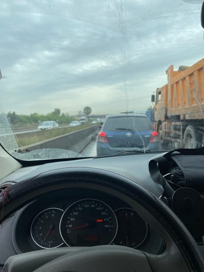 The image shows a view from inside a vehicle stuck in traffic in what appears to be Sidi Moussa, Algeria. A large dump truck is visible ahead, followed by a blue Toyota.  The vehicle's dashboard and speedometer are visible, showing a speed of 0 km/h. The weather appears overcast.