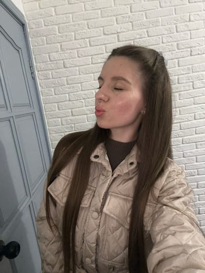 A woman is seen from a slightly elevated, angled perspective indoors. The background includes a white brick wall and a blue-grey paneled door or wall feature. She is wearing a tan, quilted jacket and a dark top underneath. Her brown hair is partially pulled back. She has her eyes closed and is puckering her lips in a kissing gesture. The photo was not taken in the city of Klin, Russia; the image does not provide any location-specific information.