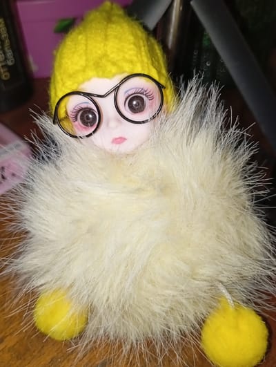 This is a close-up, eye-level, indoor photograph of a small, fluffy, yellow doll. The doll is wearing a yellow knitted hat and large, black, round-rimmed glasses. Its body is obscured by a fluffy white or cream-colored boa-like accessory, through which two yellow pom-pom feet are visible. The doll's face is partially visible, showing large, dark eyes with pinkish blush on the cheeks and pink lips. The background is out of focus and shows dark colors, possibly furniture or other objects, with some hints of purple. There is text on a dark object on the left side of the image, partially visible and in a foreign script. The image appears to have been taken in a domestic setting, possibly a home. It is not possible to determine the specific location within Рубёжин ауылдық округі, Kazakhstan, based solely on the visual information provided.
