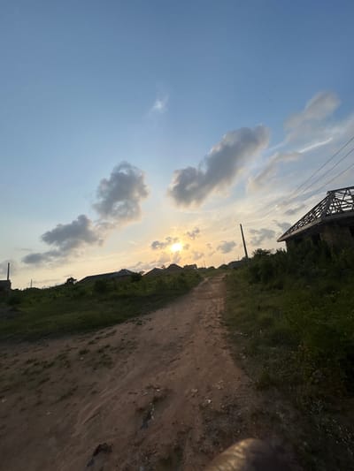 The image portrays an unpaved, reddish-brown dirt path ascending a gentle incline in Debasco, Nigeria, during late afternoon or sunset. The sky is light blue with scattered cumulus clouds, and the sun is visible as a bright, partially obscured disc near the horizon, slightly left of center. Dense green vegetation borders the path on both sides. On the right, an unfinished building with exposed concrete block walls and an incomplete wooden roof truss system is prominent, adjacent to a dark utility pole supporting overhead lines. In the mid-ground on the left, several structures with dark roofs and a concrete pillar are partially visible. No individuals, actions, or interactions are discernible. A dark, indistinct object occupies the bottom right corner of the frame.