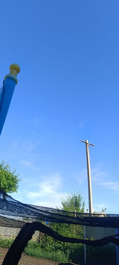 The image is an outdoor scene with a bright blue sky. In the foreground, a portion of a blue object with a yellow cap is visible on the left side, angled upwards. A black net is draped in the lower part of the frame, obscuring a view of what appears to be a garden or yard with green plants and rows of cultivated soil. In the mid-ground, a concrete utility pole stands tall against the sky. The location is specified as Khokhly, Russia. The image is taken during daylight hours.