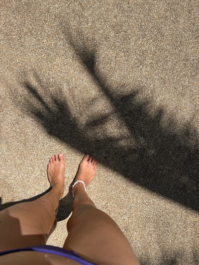 This image shows a pair of bare feet standing on a sandy surface. The sand is a light brown color and appears to be coarse, likely beach sand. A shadow of palm fronds stretches across the sand, indicating the presence of palm trees nearby and a sunny day. The feet are positioned in the lower-left portion of the image, with toes clearly visible. A white anklet with small beads is worn on the right ankle. The lower legs of a person are also visible, extending up from the feet. The environment suggests a tropical or beach setting, consistent with locations like Kathu, Thailand, known for its beaches and palm trees.