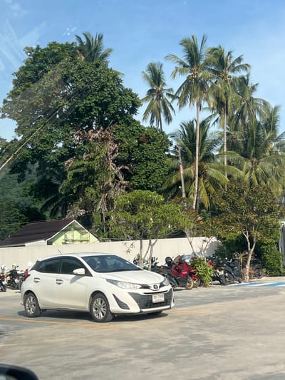 The image depicts a parking lot scene in Baan Lamai, Thailand, under a bright blue sky with scattered clouds. In the foreground, a white hatchback car is parked. To the left of the car, several motorcycles are visible. Behind the parking lot, a light green building with a dark roof is situated. The background is dominated by lush green trees, including several tall palm trees. The overall environment suggests a tropical, possibly coastal, location.