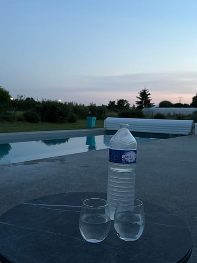 A water bottle and two empty glasses sit on a dark, round, stone-like surface in Veauchette, France. The surface appears to be an outdoor table or countertop next to a swimming pool. The pool's water reflects the surrounding trees and sky, suggesting a location near nature. Beyond the pool and trees, the sky is a gradient of light blues and pinks, indicating either dawn or dusk. The bottle is a "Cristaline" brand water bottle. No people are visible.