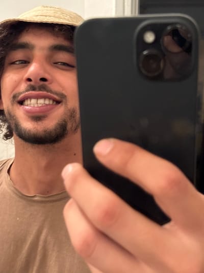 A man takes a selfie with a black iPhone. He smiles, revealing slightly crooked teeth. He wears a light-colored bucket hat and a tan t-shirt. His beard is dark and well-groomed. Part of his face is obscured by the phone, held in his hand. The phone's camera lenses and flash are visible. The background is blurry, suggesting an indoor setting.