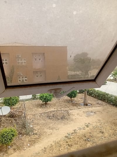 The image depicts an outdoor scene in Kwazailewa, Nigeria, viewed from an elevated position through a partially open, dusty window. In the immediate foreground, a light-colored window frame, angled upwards and to the right, is visible, featuring a white latch mechanism. The window pane is heavily marked with dust and smudges.
Below the window, the midground consists of a dry, light brown dirt area, sparsely covered with small, trimmed green bushes and scattered dark organic debris. A white satellite dish is present on the left side of this ground area. A faint wire fence line runs horizontally across the midground, and a light pole with a dark base is located towards the right.
The background is dominated by a two-story, tan-colored building with a flat roof. This building features multiple square and rectangular windows, along with several external air conditioning units mounted on its facade. Faint dark text, possibly "Tethered," is discernible on the building's front. To the right of the building, the distant environment appears hazy or dusty, obscuring clear details of trees and other structures. No individuals are present in the scene.