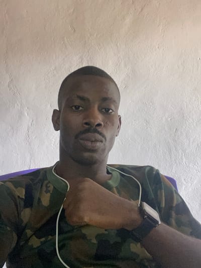 An adult male subject is depicted in a close-up, chest-up shot, seated indoors against a light-colored, textured wall. The individual has dark skin, short dark hair, a mustache, and a thin beard. Notable features include two linear scars on the subject's right cheek. He is wearing a green, brown, and black camouflage-patterned t-shirt with a white trim around the collar. White wired earphones are draped around his neck and over the t-shirt. On his left wrist, a dark-strapped digital watch with a silver-toned rectangular face is visible. The subject's left arm is raised, with his hand forming a loose fist that rests against his upper chest area. His gaze is directed towards the camera with a neutral expression. A purple object, possibly the back of a chair, is partially visible on the right side of the frame behind the subject. The scene is located in Kado, Nigeria.