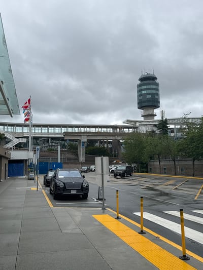 This media file captures a cloudy day scene at an airport or transportation hub in Richmond, Canada. The prominent subject is the airport control tower, a cylindrical structure with a checkered facade, visible against a grey, overcast sky. Adjacent to the tower is a multi-story building with extensive glass windows, suggesting a terminal or office complex.

Several black vehicles, including a luxury sedan with prominent headlights and grille, are parked or driving on a wet asphalt surface. The ground is marked with white and yellow lines, indicating parking or traffic lanes, and a prominent crosswalk with yellow tactile paving. Yellow bollards are strategically placed along the edge of the walkway.

In the foreground, several flags are displayed on poles, including what appear to be national flags. Some construction or temporary structures, possibly modular buildings or barriers, are also visible. The overall atmosphere is subdued due to the weather, with no clear indications of specific activity or emotions beyond the general functionality of a transportation area.

Visible text includes "VALET" on a sign pointing to the right, and a license plate on the black sedan which reads "192-SRI". The scene appears to be set during daytime, given the lighting conditions.
