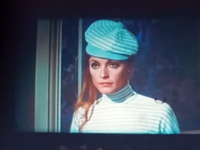 This is a still image, likely from a film or video, showing a close-up of a woman wearing a light blue and white striped cap and a matching striped turtleneck shirt. She has long, reddish-brown hair and is looking slightly to her left. Her makeup is noticeable, with dark eyeliner and mascara. The background is dark and out of focus, with some blurred blue and white shapes that could suggest an interior setting, perhaps near a window or a light source. There are some light-colored vertical elements on the left side of the frame, which might be part of a doorway or window frame. The image has a slightly grainy texture and some imperfections, suggesting it may be an older recording or has been transferred from a physical medium. No text is visible in the image. The overall mood is somewhat mysterious or dramatic due to the lighting and the woman's gaze.