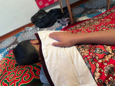 A person is lying down, seen from above and at an angle. The person is covered by a white blanket on their torso and a red and gold patterned fabric on their side. The person's head is on a pillow with a red and green swirling pattern. Their dark hair is visible. A person's hand is resting on the white blanket. The scene appears to be indoors, with a floor covered by patterned rugs. In the background, there are piles of dark clothing and a black bag. Part of a wooden chair is visible. The location is specified as Тайгараев, Kyrgyzstan.