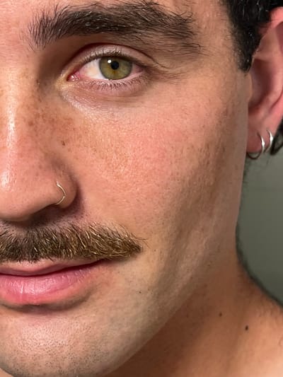 The image shows a close-up of a man's face. The man is facing the camera, and the image is cropped to include only the right side of his face, from his forehead to his neck. He has short brown hair, brown eyebrows, a mustache, and a small nose piercing. His right eye is open and looking directly at the camera, and his left eye is closed. He has light skin with freckles, and the right side of his face is illuminated, while the left side of his face is in shadow. The man is wearing a silver hoop earring in his right ear. The image was taken indoors with artificial lighting. The location is not possible to determine from the image.