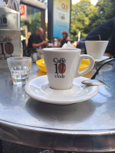 A medium shot captures a table at an outdoor cafe, likely in Turin, Italy, with a focus on a small white espresso cup and saucer in the foreground. The cup, bearing the branding "Caffè 10 elode" and a coffee bean logo, sits on its saucer, which is placed on a reflective metal table. A spoon with a residue of dark liquid rests on the saucer. To the left of the espresso cup, a small clear glass with a liquid, possibly water, is visible. In the background, out of focus, several people are seated at other tables. One man, wearing a dark t-shirt, is seated at a table, and further back, another person with reddish hair is also visible. Other cafe patrons and the blurred surroundings suggest a public, urban setting. The lighting indicates it is daytime, with natural light filtering through trees, creating a slightly dappled effect.