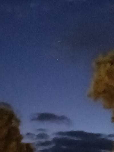 The image shows a twilight sky with two bright celestial objects, likely stars or planets, positioned vertically in the upper portion of the frame. The sky is a deep blue, transitioning to lighter shades towards the horizon. In the lower half of the image, there are blurred silhouettes of trees, one on the left with dark foliage and another on the right with yellowish-brown, possibly autumn foliage. Clouds are visible as dark smudges in the lower sky. The scene appears to be an outdoor setting, potentially a park or residential area on the outskirts of Netanya, Israel, given the presence of trees and the sky conditions often observed during dusk or dawn. There are no people or visible human activity.