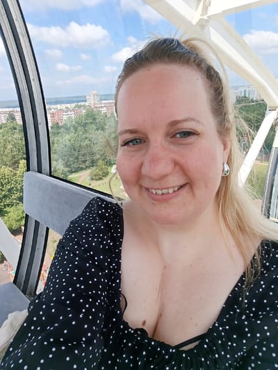 A fair-skinned Caucasian woman with blonde hair tied in a ponytail smiles for a selfie. She is wearing a black dress with white polka dots and silver hoop earrings. She is seated in what appears to be a Ferris wheel cabin, with a gray seat visible to her left. Outside the cabin's windows, a cityscape with trees and buildings is visible under a partly cloudy sky. The Ferris wheel structure itself, painted white, is also partially visible. The Ferris wheel is located in Naberezhnye Chelny, Russia.
