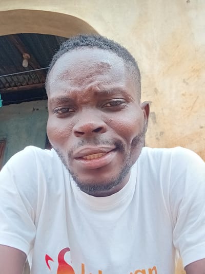 This is a selfie of a Black male in his late 20s to early 30s, wearing a white t-shirt. He has dark, curly hair, a dark complexion, and is clean-shaven with a beard. The man is looking directly at the camera, with a slight smile showing his teeth. His face is the primary focus of the image, with the background blurred. The background appears to be an outdoor setting, possibly in Ikorodu, Nigeria, with a textured, light-colored wall behind him and some corrugated metal roofing visible to the left. The lighting suggests it is daytime.