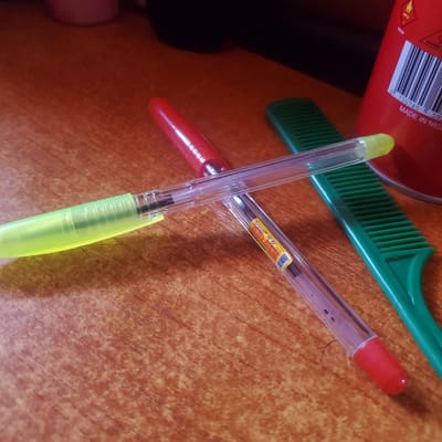 This image shows a close-up of two pens and a green comb resting on a wooden surface. One pen has a yellow-green barrel and a transparent cap, with its tip pointing downwards. The other pen has a transparent barrel and a red cap, with its tip pointing upwards. Both pens are lying on top of the green comb, which is positioned diagonally across the frame.

A red object with a barcode and some text is partially visible in the upper left corner of the image. The text "MADE IN NIGERIA" is clearly legible on this object. The scene appears to be an indoor setting, likely a desk or table, given the wooden texture of the surface. There are no indications of the time of day or weather conditions. The focus is on the arrangement of the stationery items, suggesting a still life composition rather than an active event.

The overall mood is neutral and focuses on the objects themselves. The lighting is somewhat diffused, casting soft shadows. The location context of Ribina East, Nigeria, is provided, which, combined with the visible text, suggests the items might be locally sourced or used in that region.