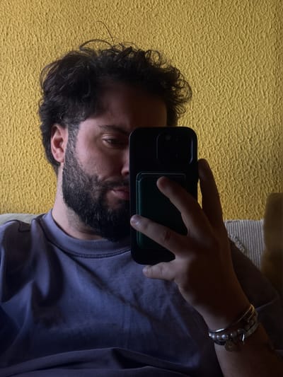 The image displays an adult male, with dark, curly hair and a dark beard and mustache, situated indoors. He wears a light purple, long-sleeved crew-neck shirt. The man's left hand holds a black smartphone, which features a dark green accessory attached to its rear and is oriented towards his face, obscuring part of his left side. His right eye is visible, directed towards the phone. The left wrist is adorned with multiple bracelets, including silver-toned bangles and a strand of pearls. The background is a textured, mustard-yellow wall. A beige striped fabric object, consistent with a cushion or part of a couch, is partially visible on the right side behind the subject. The scene is identified as being within the city of Vila Nova de Gaia, Portugal.
