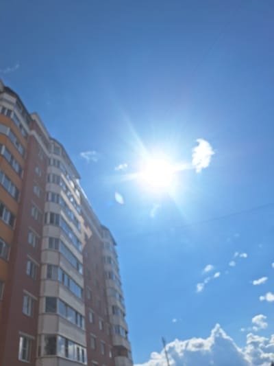 An upward-looking, slightly blurred, vertical shot captures a tall, red-brick apartment building against a bright blue sky with a prominent sun flare and scattered white clouds. The building, which appears to be located in Vologda, Russia, has numerous windows and balconies. The sun is positioned in the upper right quadrant of the image, creating a strong lens flare and illuminating the scene. There are no people visible in the frame. The overall impression is of a sunny day in an urban environment.
