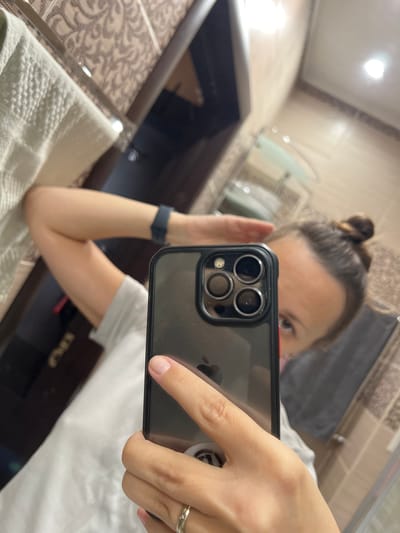 A person, with their hair in a bun, takes a selfie in a bathroom mirror. They are wearing a white t-shirt and a dark watch on their left wrist. Their right hand holds a black iPhone with a clear case, displaying the camera lenses. The background shows bathroom tiles and a towel. The image is taken in Tolyatti, Russia.