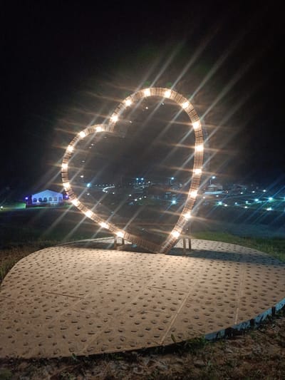 A heart-shaped sculpture illuminated by numerous lights stands prominently in the foreground of a nighttime scene in Zarubino, Russia. The sculpture is made of segmented panels, each containing a light, creating a radiant effect. It is mounted on a circular platform paved with square tiles, each featuring embedded pebbles. Beyond the sculpture, the background reveals a dimly lit town with scattered lights from buildings and streetlights. In the distance, a white building with blue accents is visible on the left side. The overall environment suggests an outdoor public space, possibly a park or recreational area, decorated for an evening display.