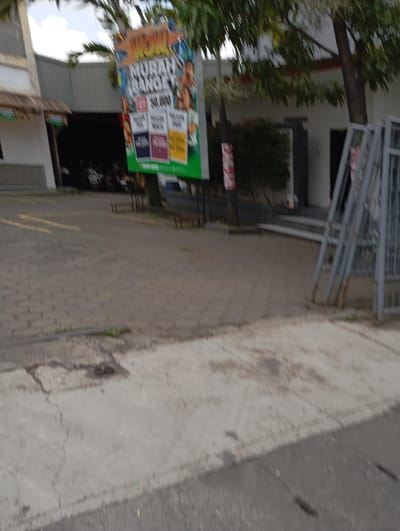 The scene is an outdoor, paved area in Katapang, Indonesia, located at coordinates -6.988 latitude and 107.558 longitude. The environment features a brick walkway, a concrete curb, and a paved street in the foreground. A large, white building with a large billboard advertising "Wow! Murah Banget" (Wow! Very Cheap) is on the left side of the image. Several parked motorcycles can be seen in the distance, partially obscured by trees and the building. A large tree with green leaves is in the right foreground. A metallic gate, leaning against the building, is partially visible in the right side of the frame. The area is empty of people.