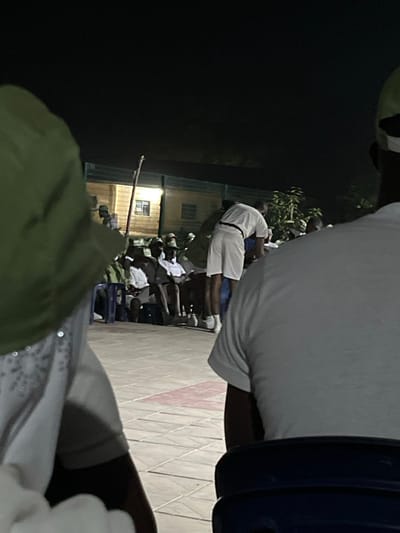 An outdoor gathering at night features many people, mainly in white, some seated, in front of a lit building. A person in a green hat is in the foreground. A man in a white shirt is prominent. Tiles with a red design are partially visible.