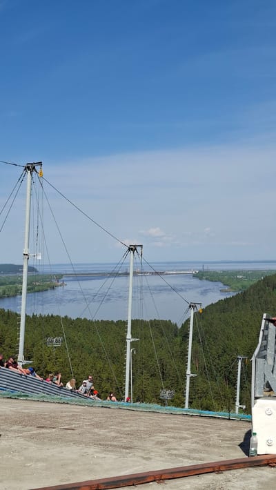 View from a high structure in Прикамский, Russia, showing an electrical transmission line tower and transmission lines, the Kama River, and an expanse of evergreen forest on the shoreline. The blue sky is visible to the left. People are standing on a gray, ribbed metal structure. The edge of a concrete structure with a rusted metal edge is visible to the right.