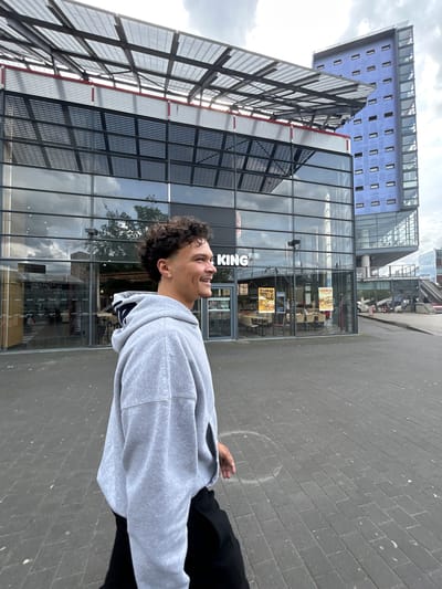 An outdoor, portrait-oriented photograph depicts a young man on a paved urban sidewalk, looking upwards and smiling or laughing. He has dark, curly hair, light stubble, and wears a light grey hooded sweatshirt and dark trousers. A small golden earring is visible in his left ear. His right hand is seen at his side.

Behind the man is a modern building with a large glass facade, reflecting the cloudy sky and trees. Part of the "BURGER KING" logo is clearly visible on the glass, specifically "KING". Inside the restaurant, through the glass, several tables and chairs are visible. Posters displayed inside include one with the word "FRENCH" and another partially obscured, possibly reading "KING WATILLA" or "KING WRAPS", advertising menu items.

Further in the background, another distinct building with a light blue or purple facade and numerous small rectangular windows stands. The ground is paved with grey rectangular bricks. The sky is overcast with visible clouds, suggesting daytime conditions. The man's upward gaze and cheerful expression convey a positive emotion.