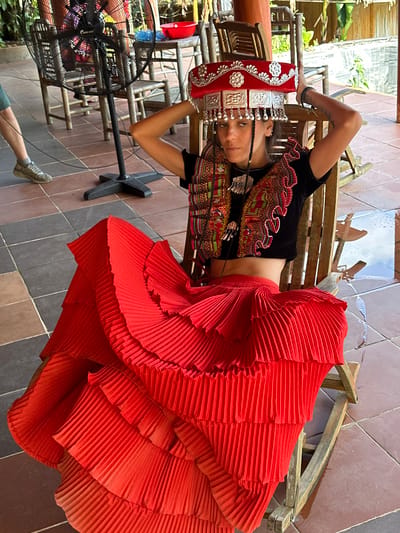 A young woman is depicted lying on her back on a wooden bench, gazing directly at the camera. She is adorned in a striking traditional costume set against a tiled outdoor or semi-outdoor setting in Thông Nguyên Commune, Vietnam.

Her attire features a voluminous, multi-tiered pleated red skirt that cascades around her, and a black short-sleeved top rich with vibrant, geometric embroidery in red, green, and black patterns. A prominent, elaborate headpiece rests on her head, crafted from red fabric decorated with silver floral motifs and multiple silver plaques and dangling ornaments that frame her face. Her hair is styled in braids on either side of her face. Her pose is relaxed, with one arm bent behind her head and the other resting on the bench, lightly holding her headpiece.

The environment includes a floor covered in various shades of brown and grey square tiles. In the background, other simple wooden benches are visible. To the lower left, a large black standing fan, with the text "OCEANIC" visible on its grille, suggests warm conditions. The leg and shoe of another individual are partially visible near the fan in the foreground. The bright natural lighting indicates it is daytime. The scene conveys a cultural portrait or photo shoot, highlighting traditional Vietnamese ethnic attire in a casual outdoor setting.