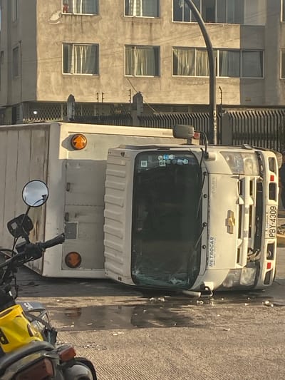 The image depicts a traffic accident scene in Quito, Ecuador. A white truck, with the branding "METROCAR," has overturned onto its side. The truck's cab is on the right side of the image, and its rear cargo container is on the left. The license plate of the truck is visible and reads "PBY-4309." The truck is positioned on a textured, gray road surface which appears wet in places, with debris scattered around it. In the foreground, a portion of a motorcycle is visible, with a yellow fender. In the background, a multi-story building with a concrete facade and several windows is visible. Some of the windows have curtains drawn. A lamppost or utility pole is present near the overturned truck. The overall lighting suggests it is daytime, with sunlight casting shadows.