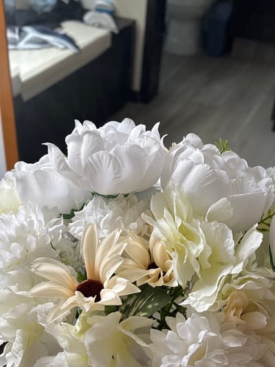 A close-up shot shows an arrangement of artificial white and beige flowers. The focus is on the floral details, with several types of flowers intermixed, including what appear to be peonies and daisies. Some of the flowers have dark brown centers. A blurry reflection of a television and a white object is visible in the dark background on the left side of the frame. No specific information can be derived about the location of Santa Clara Ocoyucan, Mexico, or the people, subjects, objects, actions, or interactions within the environment, other than the flowers in the foreground.