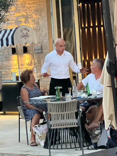 The image depicts an outdoor seating area of a restaurant or cafe in Đenovići, Montenegro. A male server, wearing a white collared shirt and black pants, is interacting with two patrons seated at a round table. The server is holding a white cup and gesturing with his right hand.
To the left of the server, a woman with short, curly brown hair is seated at the table, wearing a black and white striped dress. She is looking towards the server.
To the right of the server, a man is seated at the table, wearing a white polo shirt. He is also looking towards the server and appears to be speaking.
The table is set with glasses, bottles of water and what appears to be a bottle of alcohol. There are also cigarettes on the table.
In the background, there is a building with stone walls and vertical lighting fixtures. A striped awning with the words "ICE CREAM" is visible on the left side of the frame. The overall lighting suggests it is late afternoon or early evening.