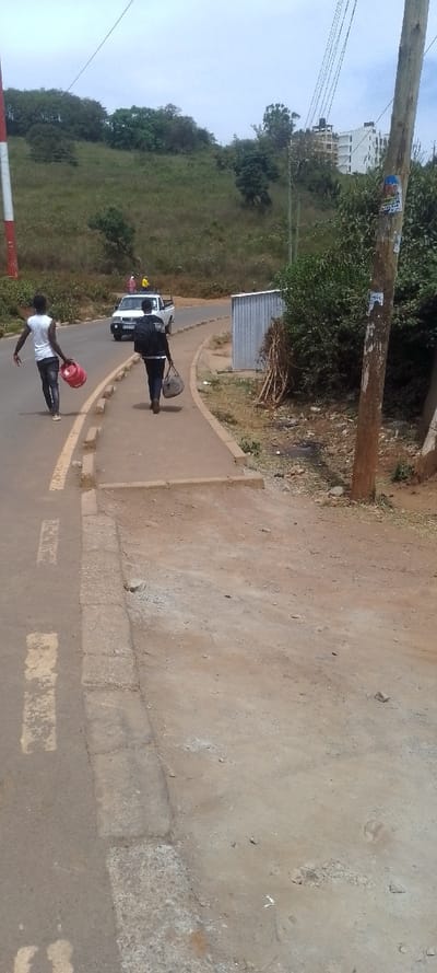 This image depicts an outdoor daytime scene in Ngong ward, Kenya, featuring a paved road, an adjacent unpaved path, and a hilly, vegetated background with distant structures under a cloudy sky.
In the foreground, a two-lane asphalt road runs diagonally from bottom-left to mid-right, marked with faded yellow solid and dashed lines on its left edge. A raised concrete curb separates the road from a reddish-brown dirt path on the right.
Two individuals are walking away from the camera. The first, on the left side of the road, is a male wearing a white sleeveless shirt and dark pants, carrying a red propane gas cylinder in his right hand. The second, on the dirt path, is a male wearing a dark long-sleeved shirt or jacket, dark pants, and a dark backpack, carrying a grey duffel bag in his left hand.
In the mid-ground, a white pickup truck, possibly a Nissan model, is visible on the road facing away, with a ladder or similar object on its roof rack. Its license plate partially reads "K_Z 791_". Two additional indistinct figures are near the truck. To the right of the dirt path, a corrugated metal structure (possibly a shed) is present, along with dense green and brown shrubbery. A weathered wooden utility pole stands on the far right, supporting multiple overhead power lines and displaying torn posters.
The background consists of a prominent green-brown hill covered in various trees and sparse vegetation. On the far right hill, multiple stories of a white building complex are visible. The sky is overcast with a uniform grey tone. A red and white striped pole, potentially a communication mast, is partially visible on the far left of the image.