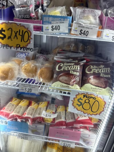 This image displays the interior of a brightly lit refrigerated display case in a store, likely an Asian grocery store given the product selection and languages. The shelves are stocked with various packaged food items, predominantly Korean and some with Spanish labels, indicating an international market setting in San Andrés Cholula, Mexico.
The top shelves feature multiple packages of "Samlip Cream Bun Chocolate" (초코생크림빵), described as a "Soft Brioche Bun" with "Rich Flavour Cream," priced with a "PROMO $80" sign. Below these, there are several individual cream-filled buns (likely "BOLLOS RELLENOS DE CREMA CHOCOLATE O QUESO") in clear plastic, priced at "$20" each.
On the left side, there are "HOTISSUE STRAWBERRY CREAM DESSERT TTEOK" (딸기크림찹쌀떡) priced at "$40" for a single piece, and a "BEBIDAS RELLENO CREMA" (cream-filled drink) also for "$40". A "Super Promo $2x40" sign is visible near a product with the text "쿠쿠돼지". Larger boxes of "HOTISSUE CREAM DESSERT TTEOK 320GR" (크림찹쌀떡) are priced at "$320".
The right side of the case holds stacks of "CASTELLA MILK CAKE" (카스테라), with a "MILK CAKE VAINILLA" variant priced at "$70". Many products feature the "K-Food" logo, and some have visible expiration dates like "2027.05.29까지" and "2027.06.01까지". The overall scene suggests a well-stocked section for frozen or chilled desserts and snacks.