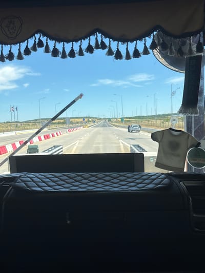 The image is taken from inside a vehicle, likely a truck, looking out through the windshield. The perspective suggests the vehicle is stationary at what appears to be a border crossing or toll plaza on a highway. The road ahead is wide, with multiple lanes separated by barriers. In the distance, the road curves and disappears over a slight rise. The sky is blue with scattered white clouds.
Decorations are visible on the interior of the vehicle's windshield. A valance with tassels hangs across the top, featuring a stylized lion emblem. A hanging air freshener shaped like a t-shirt is on the right side of the windshield, and a decorative string with tassels hangs on the left. A vertical sign reading "BULGARIA" is also attached to the windshield on the right.
In the foreground, a quilted black surface, likely part of the vehicle's dashboard or seat, occupies the bottom portion of the image. A white and black striped barrier arm is partially raised on the left side, suggesting the vehicle is either waiting to proceed or has just passed through. Red and white concrete barriers line the sides of the road. Several streetlights are visible along the highway. Two cars are on the road ahead, one white and one dark.
The location is identified as being in the city of Ardhani, Greece. The scene depicts a typical highway infrastructure, possibly at an international border, with elements of personal decoration inside the vehicle.