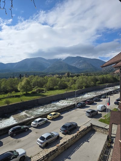 The image shows a scene in Bansko, Bulgaria. A mountain range occupies the left side of the frame, partially obscured by clouds in the sky. Below the mountains is a line of dense trees. A river runs through the frame's center, bordered by concrete banks. A road runs parallel to the river. Parked vehicles are lined up along the road. The flags of Bulgaria and neighboring countries are visible. A building with a brown roof and wooden railing is on the top right of the frame. The photo is taken from an elevated perspective, likely from a higher story of the building.