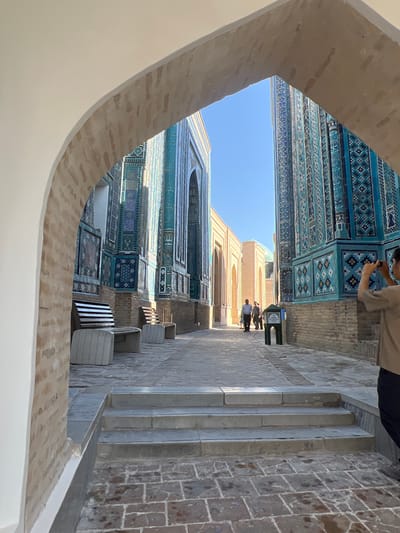 The image depicts a narrow pathway lined with ornate blue-tiled mausoleums, likely in the Shah-i-Zinda necropolis in Samarkand, Uzbekistan. The pathway is paved with cobblestones and a set of stone steps leads up from the foreground. On the left, an arched entryway with brickwork frames the scene. Benches with striped seats and stone bases are placed along the side of the pathway. In the distance, several people are walking, and one person in the foreground on the right is taking a photograph with their phone. The architecture features intricate blue and turquoise geometric patterns on the mausoleums, contrasting with the sandy-colored buildings in the background. The sky is clear and blue, indicating a sunny day.