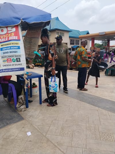 The scene is a street in Ura Nta Umuarandu, Nigeria, with various activities underway. A woman in a patterned dress stands at a POS (Point of Sale) kiosk under a blue umbrella, holding a blue device, potentially conducting a transaction. The kiosk advertises money withdrawal, deposit, send, and data purchase services for various mobile networks. A man in an olive-green shirt and cap stands nearby, observing. Another man, wearing an orange and green patterned shirt, is interacting with a young girl holding a black bowl, possibly selling goods. A gas station is visible in the background with the word "BICOZOIL" on it. Other individuals are walking in the background. The ground is paved with light-colored stone slabs. Avocados can be seen in a container beside the kiosk.
