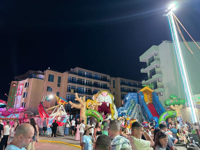 This is a nighttime photograph of an amusement park in Nesebar, Bulgaria, likely along the coast given the presence of a large building that appears to be a hotel or apartment complex. The foreground is occupied by a crowd of people, some of whom are looking towards the amusement park attractions. In the midground, there are several amusement rides and attractions, including a large inflatable slide with a dinosaur theme, a bouncy castle shaped like a tiger's head, and a pirate ship-style swing ride. Lights from the attractions illuminate the scene, contrasting with the dark night sky. Further in the background, the multi-story hotel building is visible, its windows lit from within. The overall impression is one of a lively, family-friendly entertainment area.