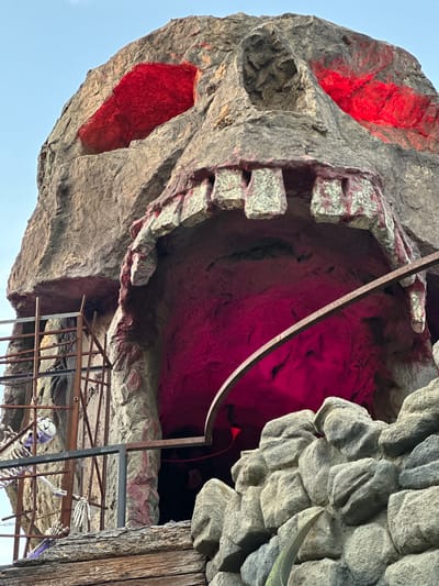 The image shows a large, artificial skull structure that appears to be a theatrical prop or attraction. The skull is made of rough, textured material resembling rock, with large, gaping openings for eyes and mouth. The eye sockets are illuminated with bright red light, and the mouth cavity is lit with a vibrant pinkish-red hue. Jagged, tooth-like protrusions line the upper jaw of the mouth.
To the left of the skull, a rusty metal scaffolding with chains is visible, and a skeletal figure hangs within the scaffolding. Below the scaffolding, there is a wooden platform with a rough-hewn texture.
The structure is set against a clear, pale blue sky. The overall impression is that of a spooky or horror-themed exhibit.
The image was taken in the Prater amusement park in Vienna, Austria, near the entrance of the "Darkride" attraction, which features a similar skull-shaped entrance.