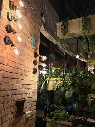 The image depicts an outdoor setting at night, likely a patio or balcony area of a restaurant or cafe. The foreground features a brick wall with several exposed filament light bulbs mounted on black bases, casting a warm glow. In the background, there are planters with lush green foliage, some cascading down over a beige structure. Large windows with a dark frame are visible behind the plants, reflecting the interior lights and greenery. Overhead, more plants and awnings can be seen, suggesting an establishment with an outdoor seating area. A light green triangular object and a dark brown shelf-like object are placed on the brick surface, along with a white electrical outlet. The overall atmosphere is cozy and inviting, indicative of a place open for evening patrons. No people are visible in the image. There is no visible text. The time of day is clearly night.