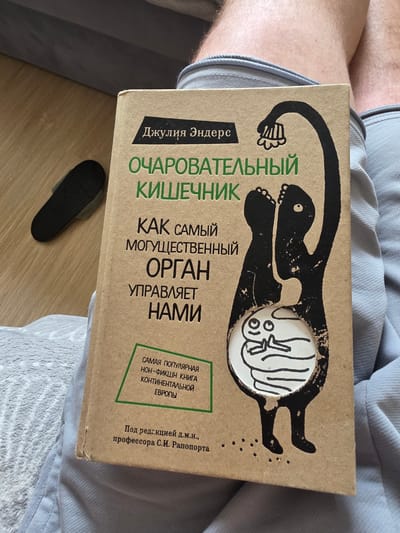 The image shows a close-up of a book titled "Charming Intestine" by Giulia Enders, held by a person whose legs are visible in the frame. The book is made of brown cardboard and features a black illustration of a stylized human figure with an open abdomen revealing a fetus-like form. The text is in Russian. The book is placed on a person's lap, with a grey blanket or fabric beneath. A black slipper is visible to the left of the book, lying on a wooden floor. The overall setting appears to be indoors, possibly a living room or bedroom. There is no indication of the city of Parshino, Russia, or any specific location within it. The scene is static and focuses on the book and its immediate surroundings.