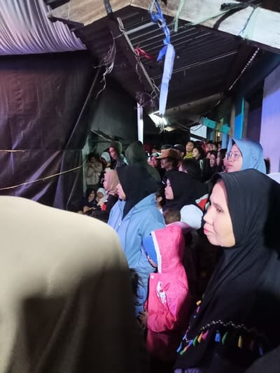 This image captures a crowded community gathering, likely an evening event, in Kecamatan Cimaung, Indonesia. The setting appears to be a semi-enclosed outdoor space, possibly a narrow alleyway or a temporary sheltered area, indicated by the dark tarp walls on the left and the corrugated metal roof structure overhead. Artificial lighting suggests it is nighttime or late evening, with a blue or purple light source illuminating the right side of the crowd.

The primary subjects are numerous people, mostly women and children, with several individuals prominently featured in the foreground and mid-ground. Many of the women are wearing hijabs, including a woman in the far right foreground with a black hijab adorned with colorful tassels, and another woman in the mid-ground wearing a light blue jacket and a black hijab. A young child in a bright pink hooded jacket and a blue hat is visible in the lower mid-right. Further back, a woman with glasses wears a light blue hijab, and men are also interspersed within the crowd.

The crowd appears to be attentive, with most people looking in the same direction, suggesting they are watching a performance, presentation, or some other focal point off-camera. Their expressions are generally calm and observant. The structures, including draped white fabric on the left and blue and white fabric strips tied and hanging from the roof, contribute to the informal, community event atmosphere. A yellow string or rope is visible across the dark tarp wall on the left. There is no discernible text visible in the image.